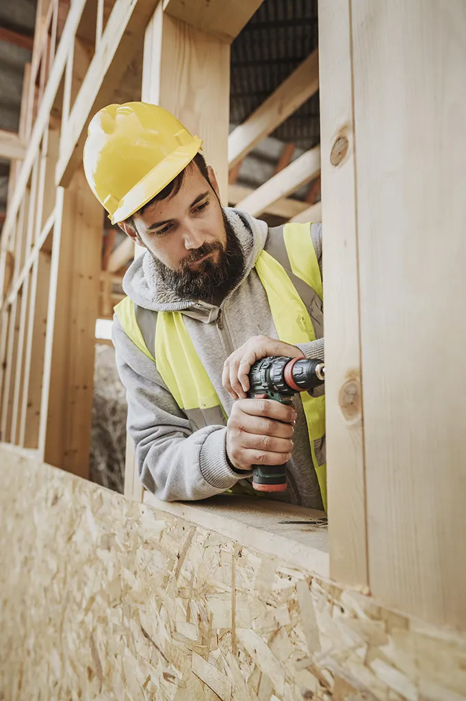 Homme portant un casque de chantier et utilisant une perceuse dans une structure en bois, illustration des interventions proches de zones électriques